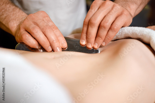 Close-up view of man s hands who massages a woman s stomach using IASTM tool for scraping muscles in spa or rehab clinic