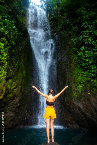 A beautiful girl spreads her arms while standing under a tropical waterfall in Costa Rica; swimming in a hidden waterfall in the rainforest; don jose waterfalls
