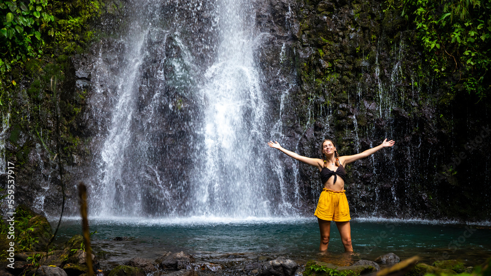Obraz premium A beautiful girl spreads her arms while standing under a tropical waterfall in Costa Rica; swimming in a hidden waterfall in the rainforest; don jose waterfalls