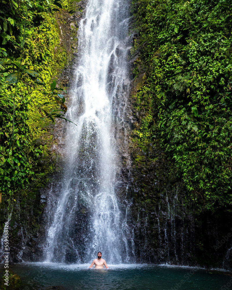 Obraz premium A relaxed man takes a refreshing bath under a tropical waterfall in Costa Rica; bathing at a hidden waterfall in the rainforest; don jose waterfalls