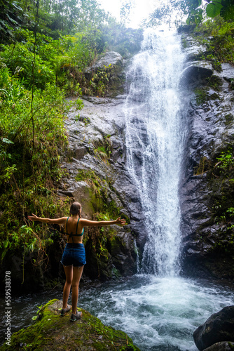 A beautiful girl spreads her arms while standing under a tropical waterfall in Costa Rica  swimming in a hidden waterfall in the rainforest