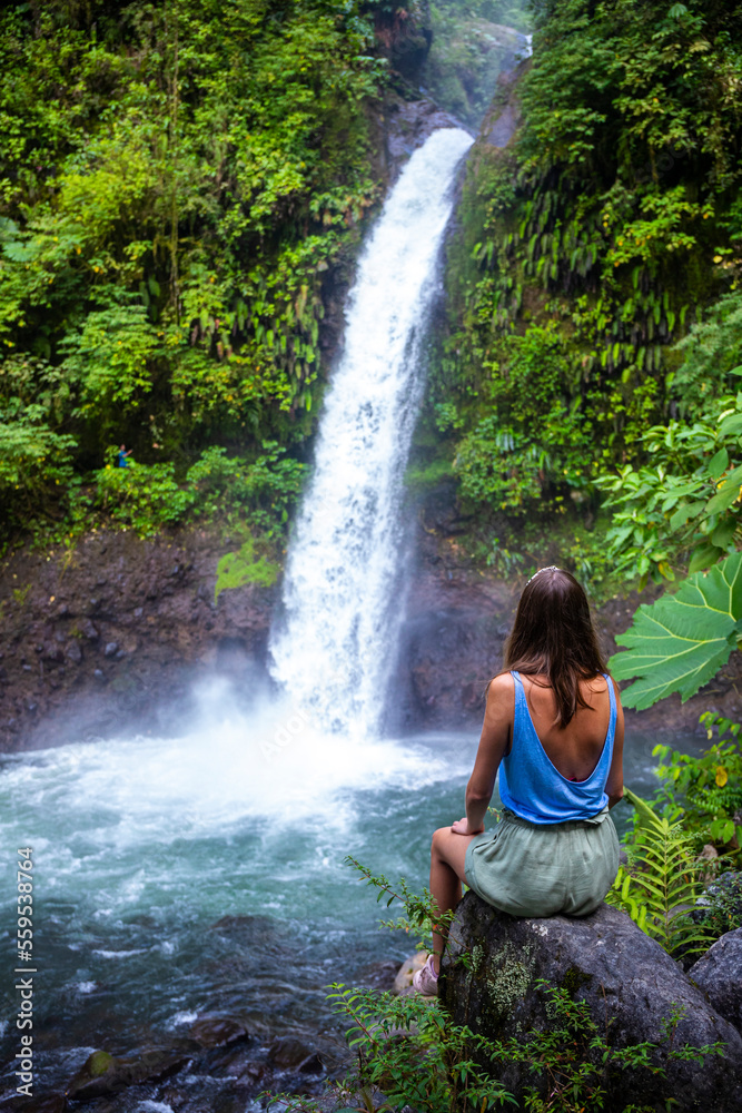 A beautiful girl sits on rocks under a powerful tropical waterfall in Costa Rica; la paz waterfall in the jungle A beautiful girl sits on rocks under a powerful tropical waterfall in Costa Rica; la paz waterfall in the jungle