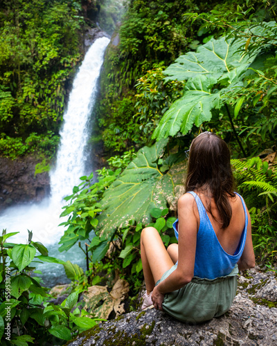 A beautiful girl sits on rocks under a powerful tropical waterfall in Costa Rica; la paz waterfall in the jungle