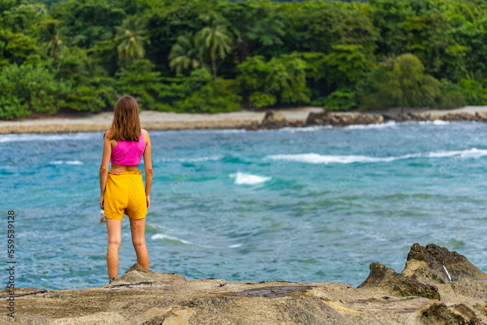 Fototapeta premium A beautiful girl in colorful clothing stands on the rocks admiring a tropical beach with palm trees; paradise beach in Costa Rica