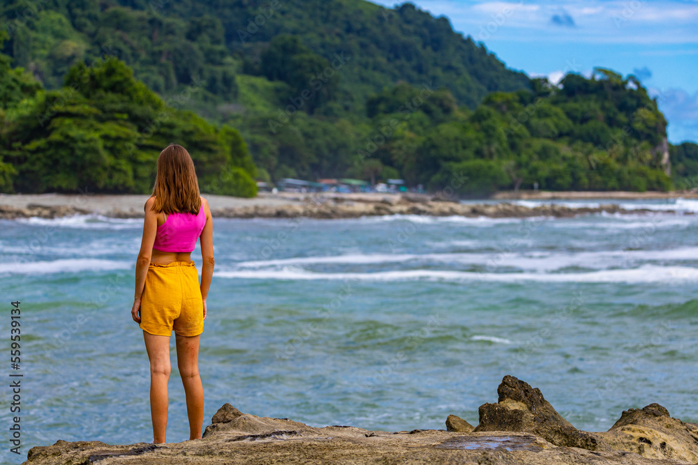 A beautiful girl in colorful clothing stands on the rocks admiring a tropical beach with palm trees; paradise beach in Costa Rica