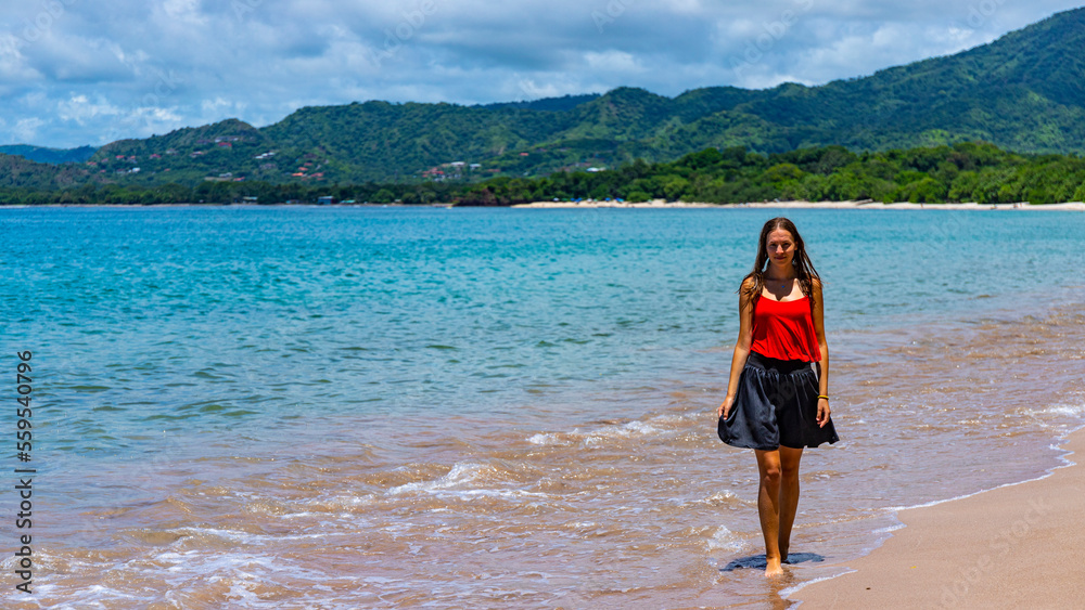 a beautiful girl in a short skirt walks on the famous conchal beach on the pacific coast of Costa Rica; a paradise beach with turquoise water and green hills in the background