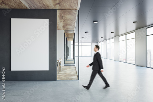 Walking businessman by blank white poster on black wall background in sunlit modern interior design office with concrete floor, meeting room with wooden decoration and city view background, mock up