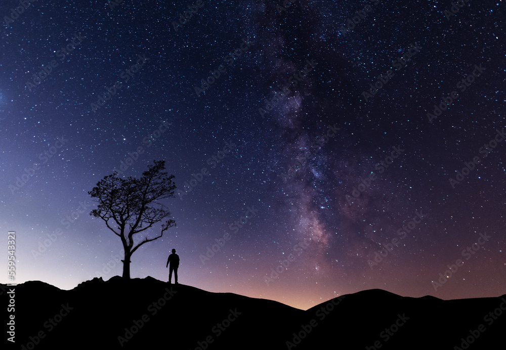 Hiker silhouette stands near the tree on the hill, behind them fantastic beautiful night sky with milky way galaxy. 