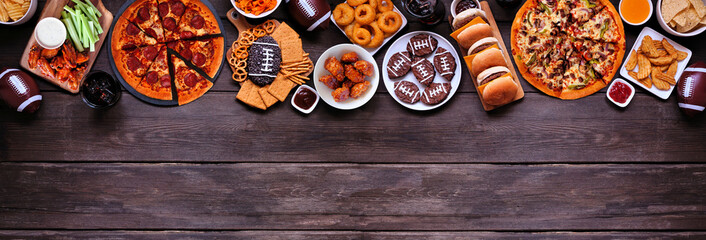 Super Bowl or football theme food top border. Pizza, hamburgers, wings, snacks and sides. Overhead view on a dark wood banner background.