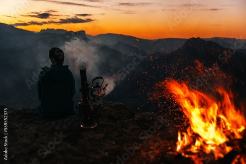 A man sits in the mountains and watches the sunset near a fire