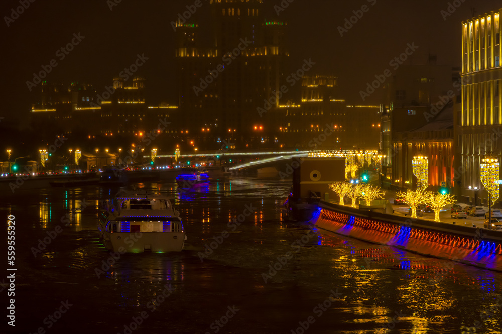 City embankment decorated with gold colored illuminated tree shaped artificial Christmas decorations at night. Tourboat on water. Selective focus. Winter holidays theme.