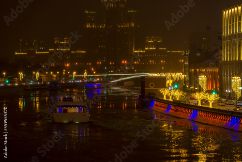 City embankment decorated with gold colored illuminated tree shaped artificial Christmas decorations at night. Tourboat on water. Selective focus. Winter holidays theme.