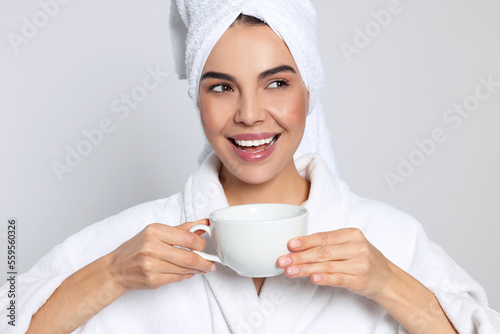 Young woman in bathrobe with towel and cup of drink on light grey background. Spa treatment