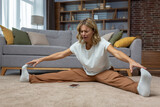 Dissatisfied and tired woman trying to stretch while sitting on floor in living room at home.