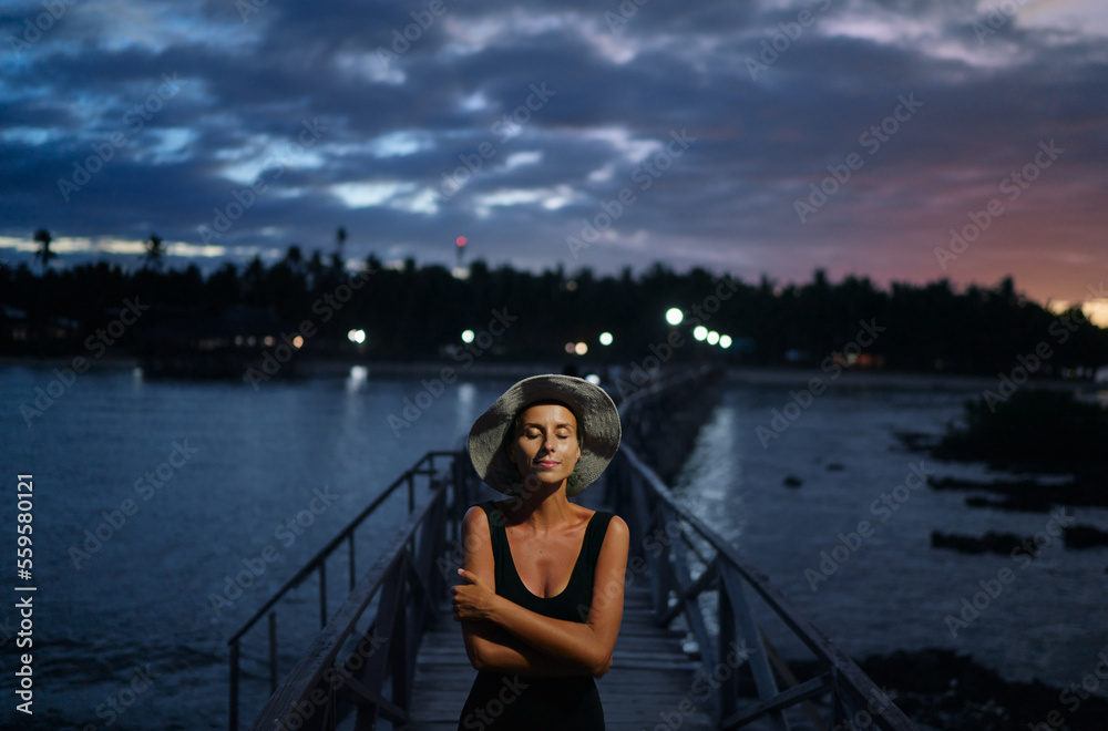 Vacation on tropical island. Young woman in hat enjoying sea view from wooden bridge terrace at night time, Siargao Philippines.
