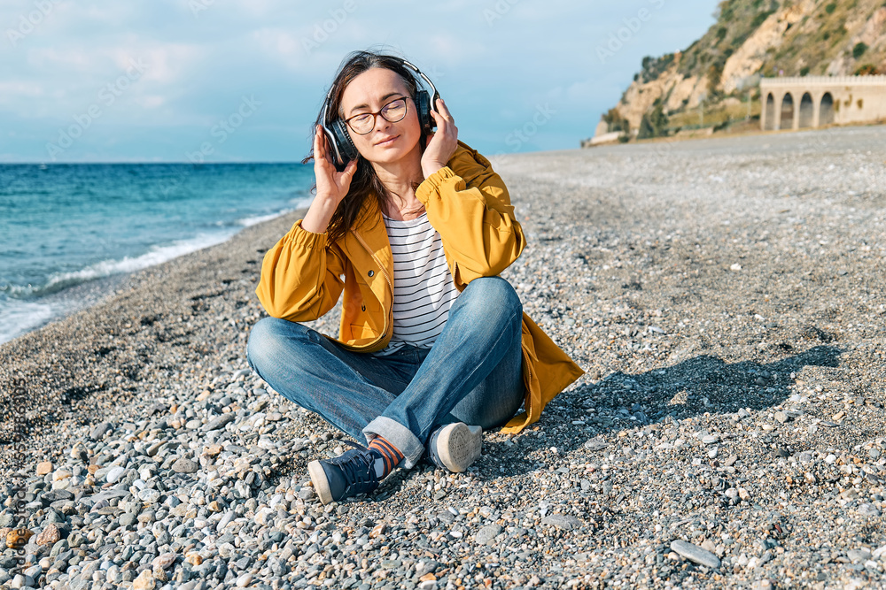 Woman with closed eyes listening music or podcast from smartphone application in headphones while sits on winter beach. Smiling middle-aged female in headphones listening music near the sea.