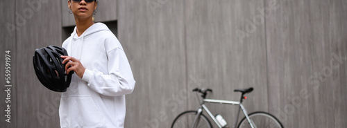 Banner shot of african american woman in hoodie holding cycling helmet on street 