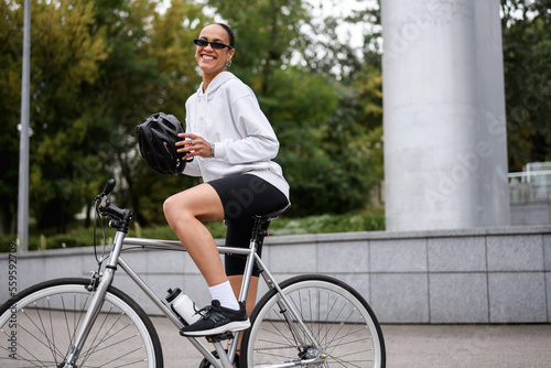 Joyful african american cyclist in sunglasses holding helmet near bike on street 