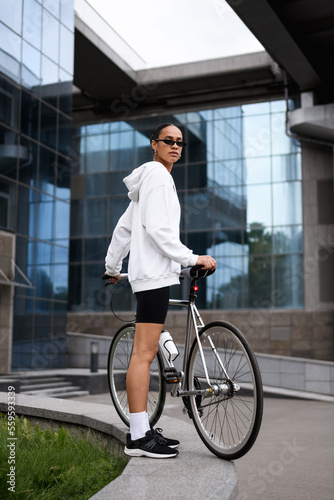 African american woman in sunglasses and cycling shorts standing near bike on street 
