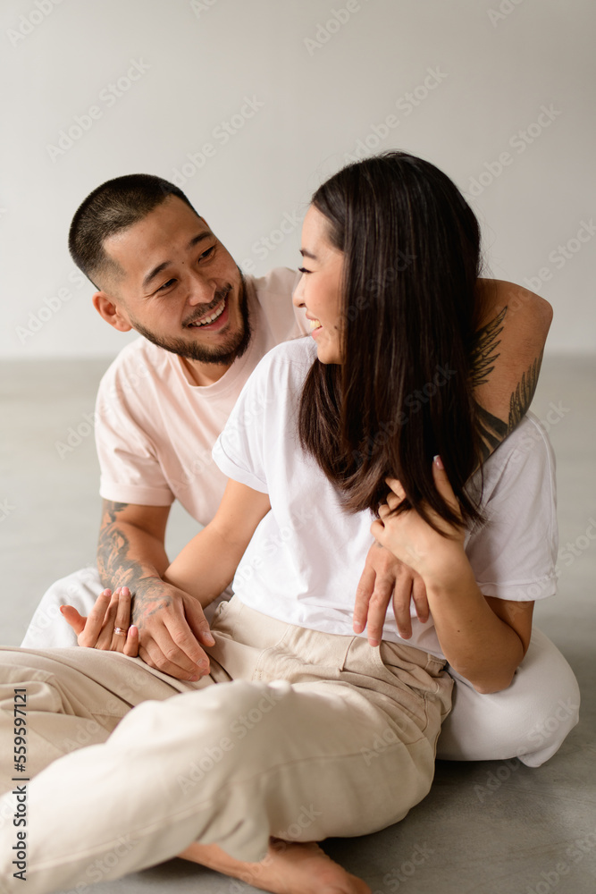 Smiling tattooed asian husband hugging young brunette wife while sitting on grey background Smiling tattooed asian husband hugging young brunette wife while sitting on grey background