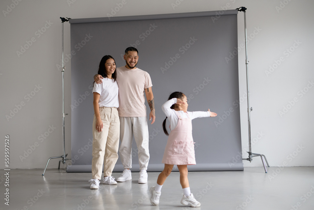 Fototapeta premium Smiling asian parents hugging while toddler daughter dancing on grey background 