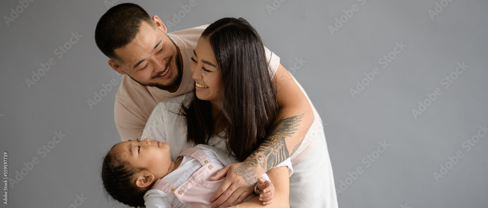 Tattooed asian man hugging daughter and smiling wife isolated on grey, banner view Tattooed asian man hugging daughter and smiling wife isolated on grey, banner view