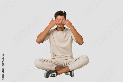 Young caucasian man sitting on the floor isolated on white background afraid covering eyes with hands.