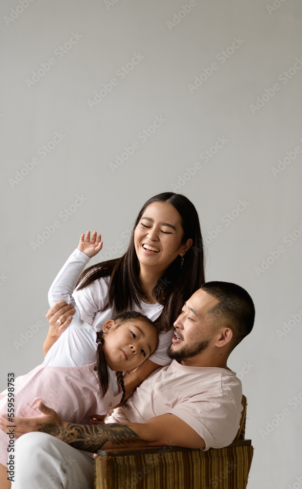 Happy young asian couple playing with toddler daughter on armchair isolated on grey background 