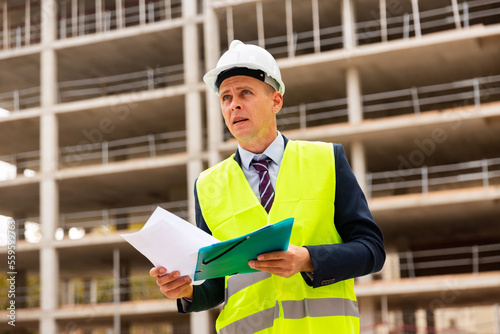 Civil engineer standing on a construction site, carefully studies important work documents © JackF