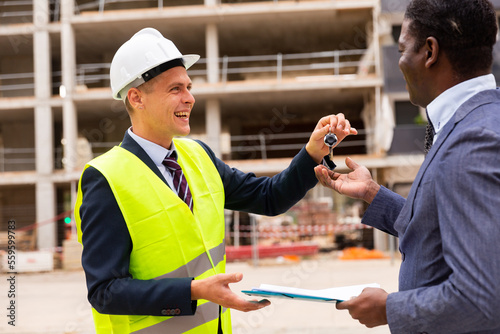 Young smiling contractor working on a construction site hands the keys to a flat in a new building to an african-american ..man customer holding a folder with documents in his hands
