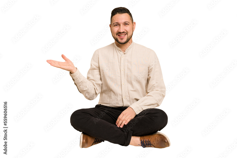 Adult latin man sitting on the floor cut out isolated showing a copy space on a palm and holding another hand on waist. Adult latin man sitting on the floor cut out isolated showing a copy space on a palm and holding another hand on waist.