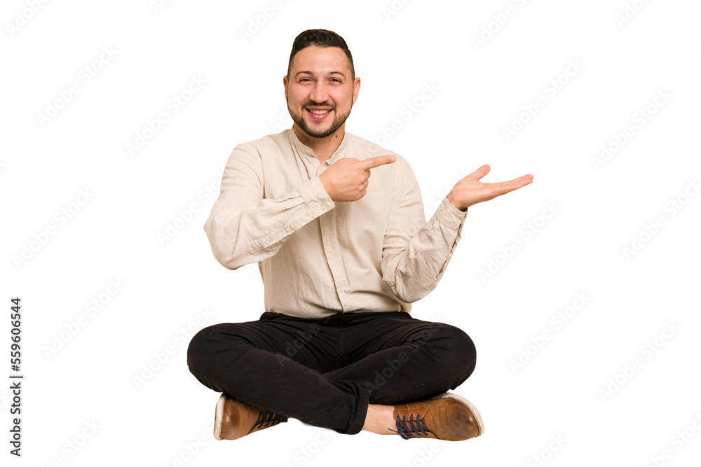 Adult latin man sitting on the floor cut out isolated excited holding a copy space on palm. Adult latin man sitting on the floor cut out isolated excited holding a copy space on palm.