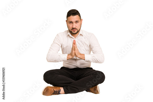 Adult latin man sitting on the floor cut out isolated praying, showing devotion, religious person looking for divine inspiration.