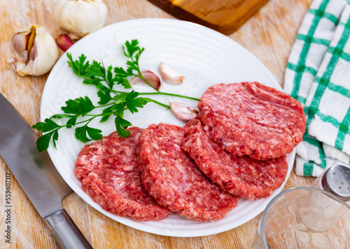 Shaped patties for burgers from raw ground beef meat with fresh parsley sprigs, spicy garlic and allspice on plate. Cooking ingredients