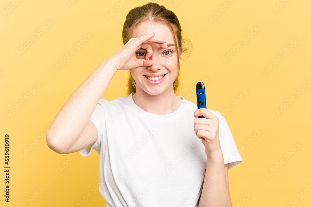 Young cute woman holding a vaper isolated on yellow background excited keeping ok gesture on eye. Young cute woman holding a vaper isolated on yellow background excited keeping ok gesture on eye.