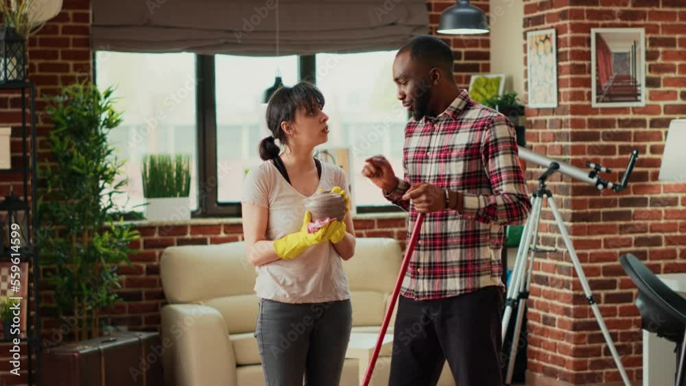 Mixed race people cleaning household with tools and equipment, husband mopping floors and wife sweeping dust off of shelves. Life partners using mop and rags to clean house. Handheld shot. Mixed race people cleaning household with tools and equipment, husband mopping floors and wife sweeping dust off of shelves. Life partners using mop and rags to clean house. Handheld shot.