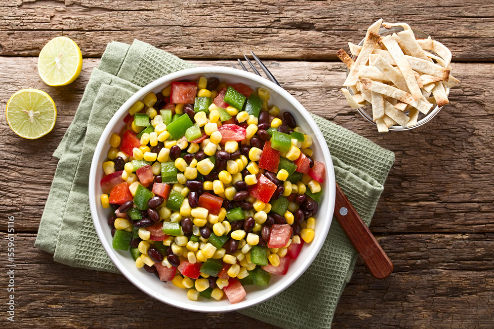 Mexican style colorful fresh vegetable salad made of beans, corn, tomato and bell pepper served in bowl, baked tortilla strips, limes and fork on the side, photographed overhead on wood - obrazy, fototapety, plakaty 