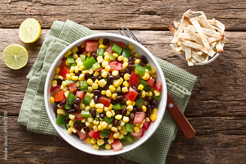Mexican style colorful fresh vegetable salad made of beans, corn, tomato and bell pepper served in bowl, baked tortilla strips, limes and fork on the side, photographed overhead on wood