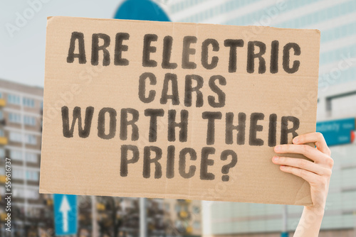 The question " Are electric cars worth their price? " is on a banner in men's hands with blurred background. Clean. Recharge. Industry. Modern. Plug. Charge. Ecological. Renewable. Drive