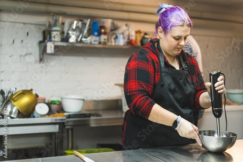 A female baker with purple hair blending products in a metal bowl on a metal kitchen counter. Blurred background . High quality photo