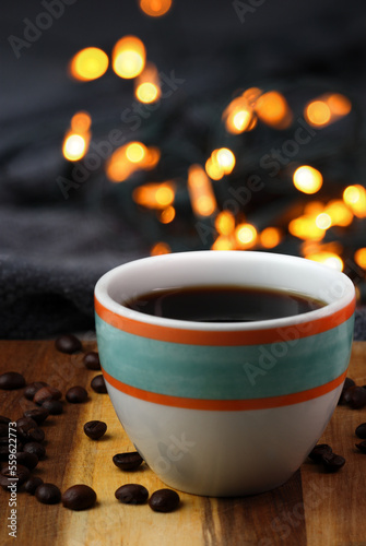 Colorful cup of black coffee with a cozy warm blanket on a wooden surface with the coffee beans and bokeh lights in the background.