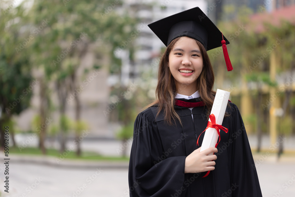 Successful graduation from university. Smiling beautiful Asian girl  university or college graduate standing with diploma over university at  background. Stock Photo