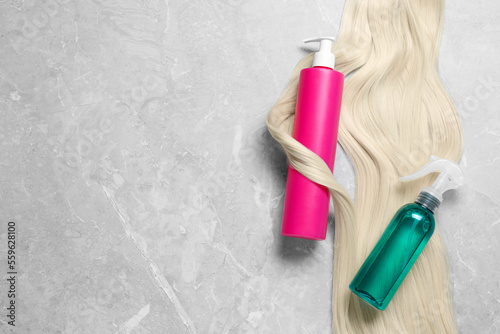 Spray bottles with thermal protection and lock of blonde hair on grey marble table, flat lay. Space for text