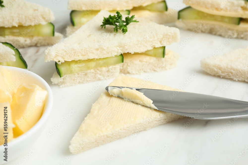 Spreading butter on tasty sandwiches with cucumber and parsley on white marble table, closeup