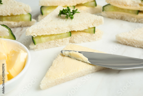 Spreading butter on tasty sandwiches with cucumber and parsley on white marble table, closeup