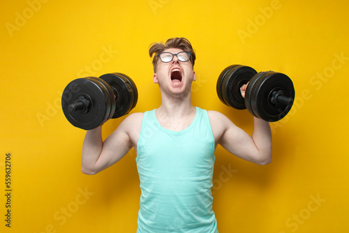 young confident guy student lifts heavy dumbbells on a pink background, motivated nerd in glasses goes in for sports