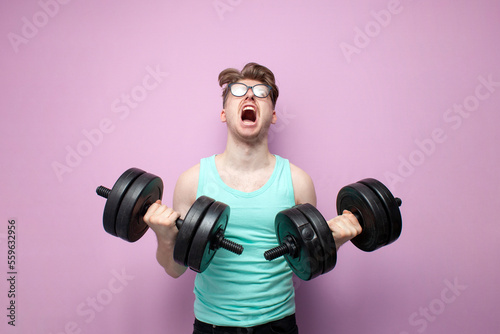 non-athletic guy in glasses holds heavy dumbbells on a pink background and trains, motivated nerd goes in for sports