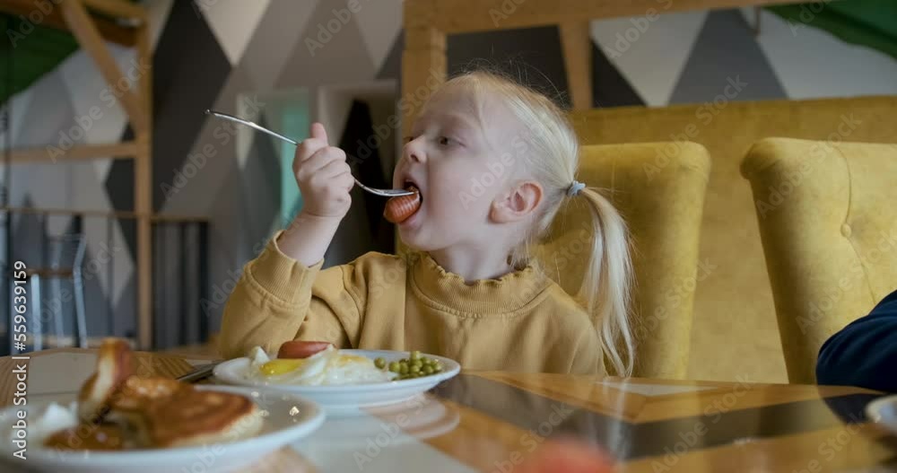Fair-haired little girl with ponytails chews sausage sitting at table in cafe. Cute child with appetite eats lunch Fair-haired little girl with ponytails chews sausage sitting at table in cafe. Cute child with appetite eats lunch