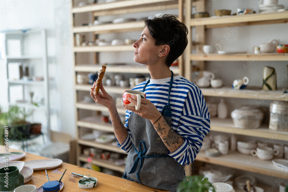 Creative Spanish woman owner of pottery studio. Female artist in apron holding handcrafted clay mug with coffee, enjoying lunch in workshop, thinking, looking at window. Ceramics master at workplace