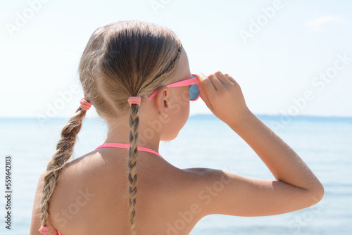 Little girl wearing sunglasses at beach on sunny day, back view